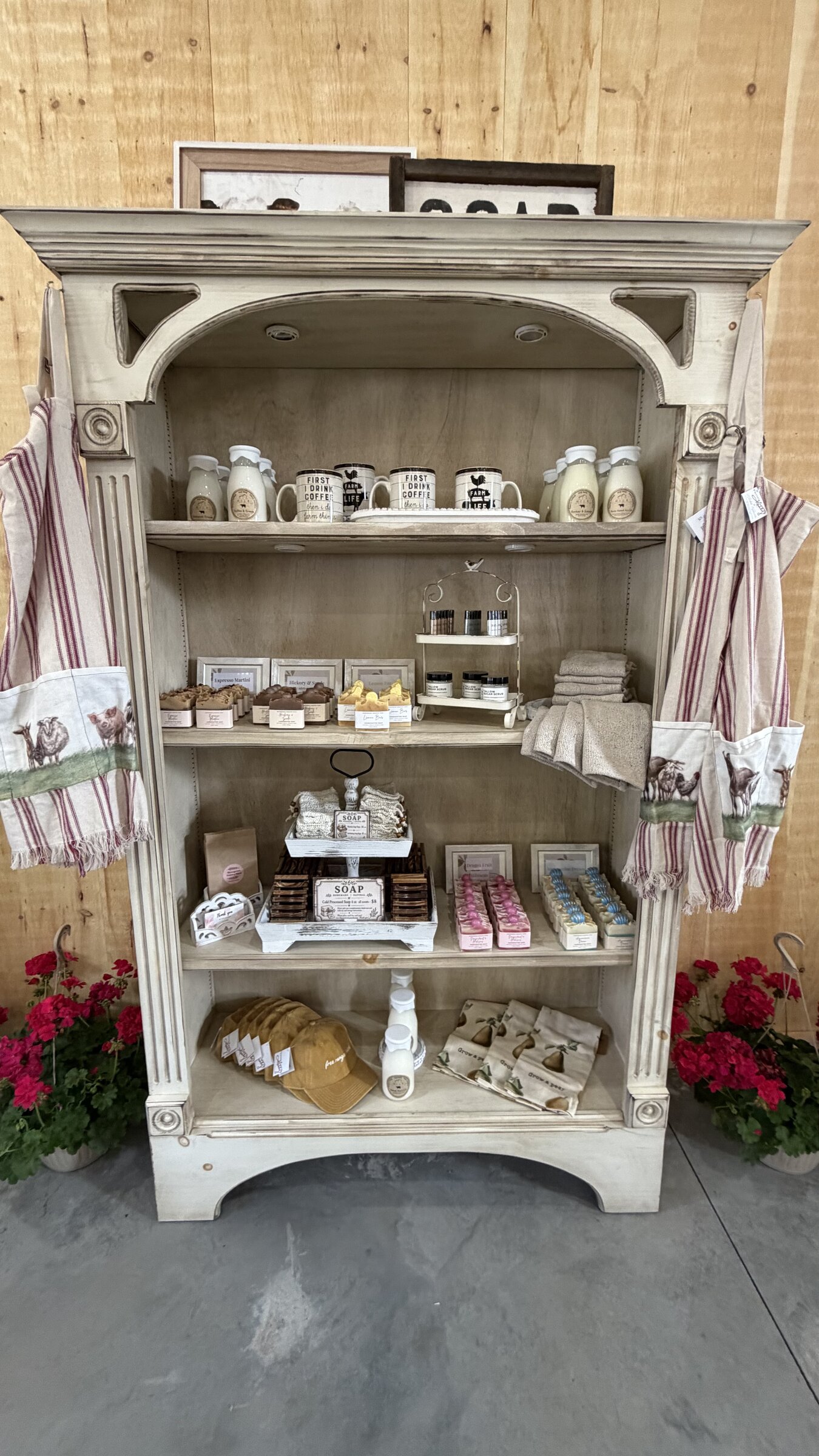 Cabinet of handmade soaps, mugs, and farm towels with red geraniums